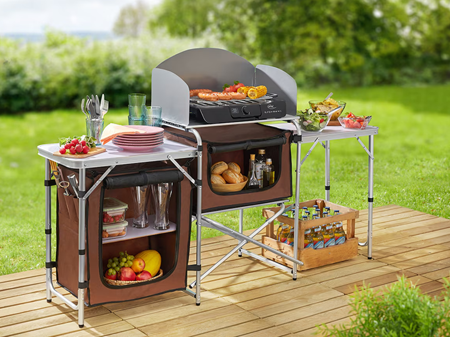 Eine mobile Außenküche mit Grill, ausgestattet mit Speisen, Getränken und Geschirr, auf einer Holzterrasse mit Blick auf einen grünen Garten.