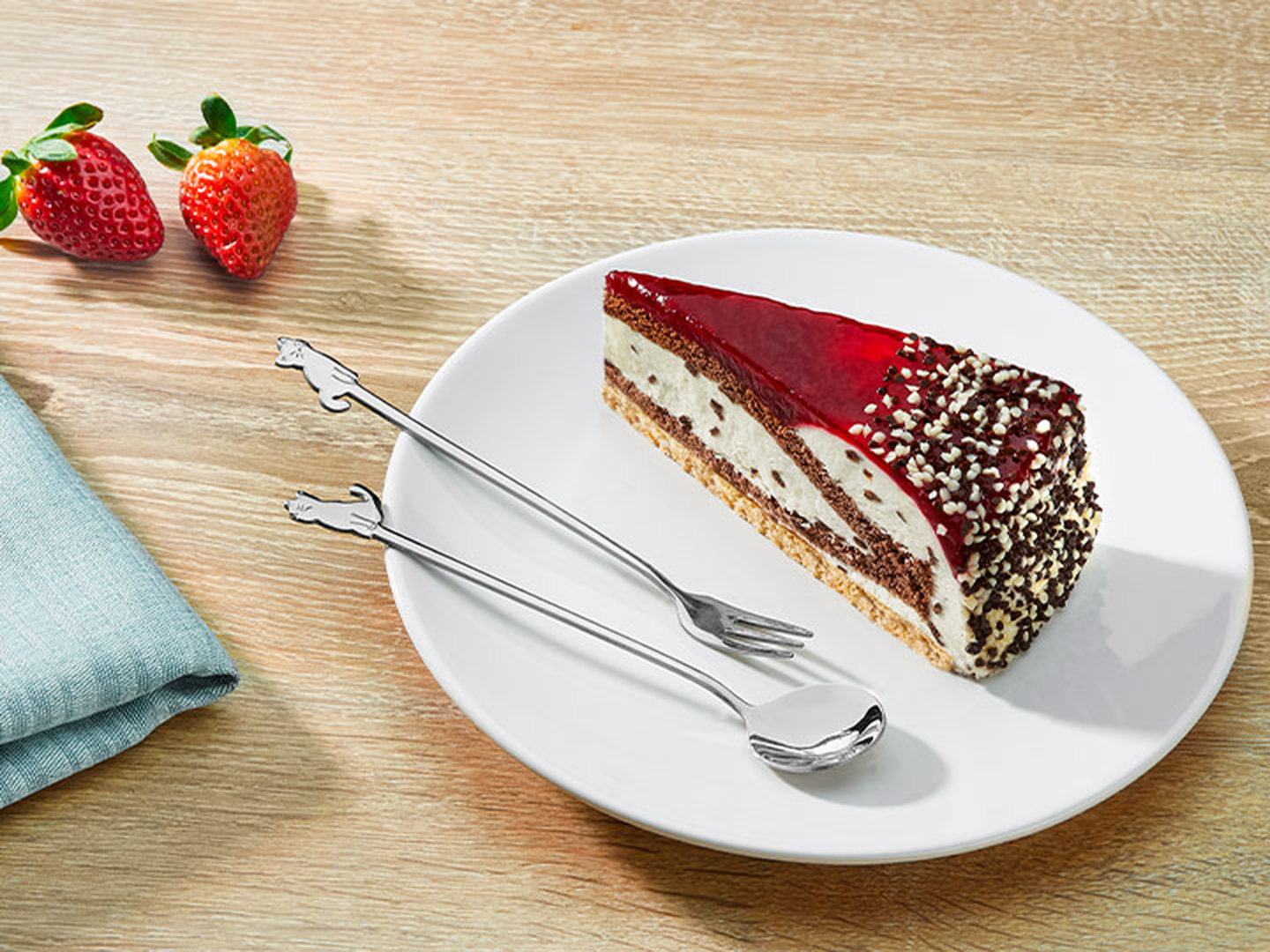 Ein Stück geschichteter Käsekuchen mit roter Glasur und Schokostreuseln auf einem weißen Teller, dazu Gabel und Löffel, daneben Erdbeeren.