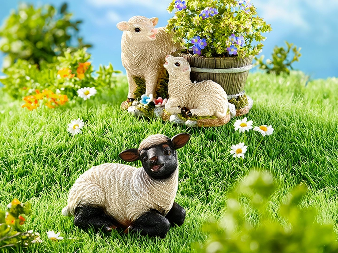 Keramische Schaffiguren im Gras, eine mit schwarzem Gesicht liegt da, zwei stehen in der Nähe eines Holzfasses mit lila Blumen, umgeben von Gänseblümchen.