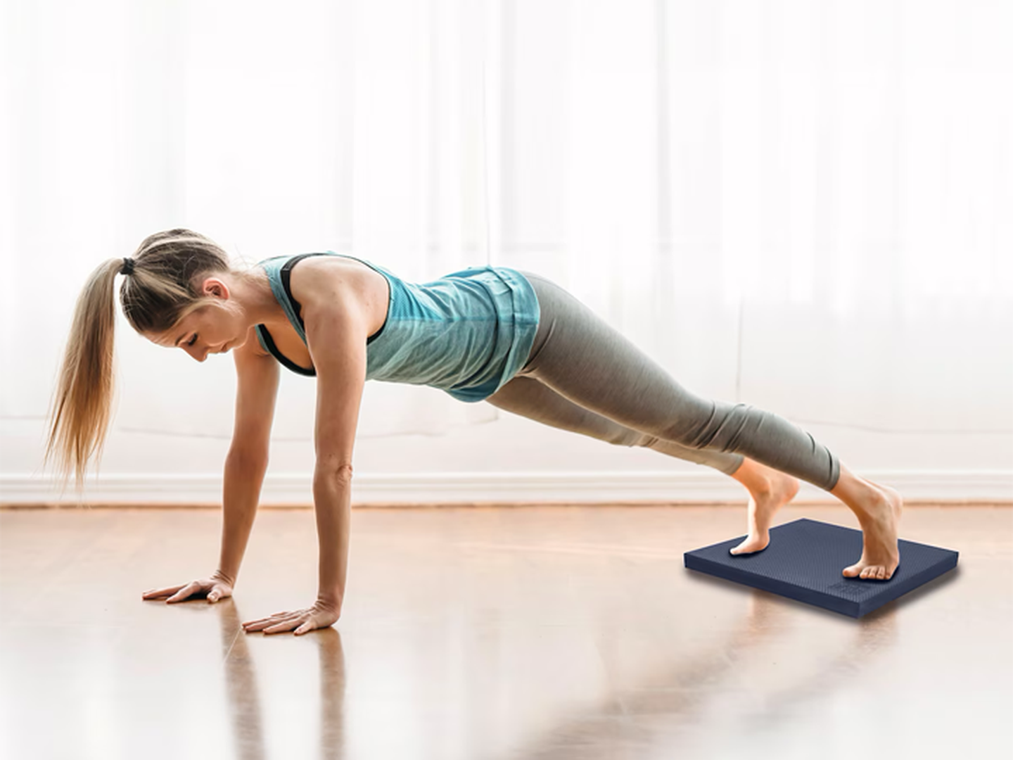 Eine Frau in Sportkleidung hält die Plank-Position, ihre Füße stehen auf einer blauen Gymnastikmatte, in einem hellen Raum mit durchsichtigen Vorhängen.