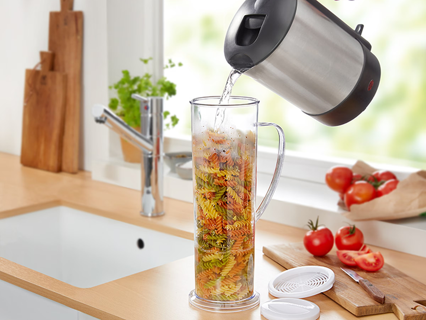 Ein Wasserkocher gießt heißes Wasser in ein hohes Glas, das mit bunten Spiralnudeln gefüllt ist; das Ganze steht auf einer hölzernen Küchenarbeitsplatte in der Nähe frischer Tomaten.