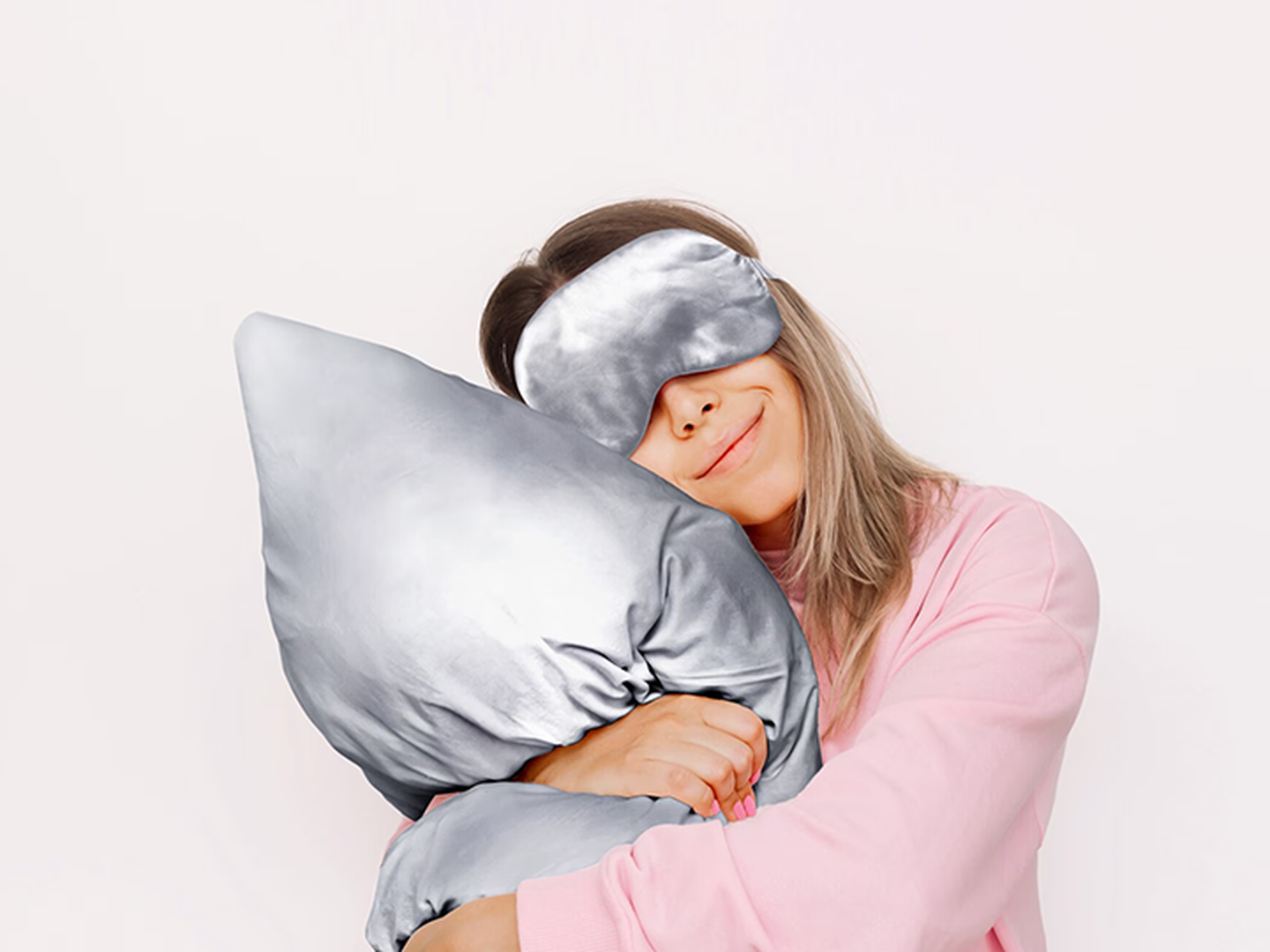 Eine Frau trägt eine graue Schlafmaske aus Satin und kuschelt ein Kissen welches auch einen Kissenbezug aus grauem Satin hat