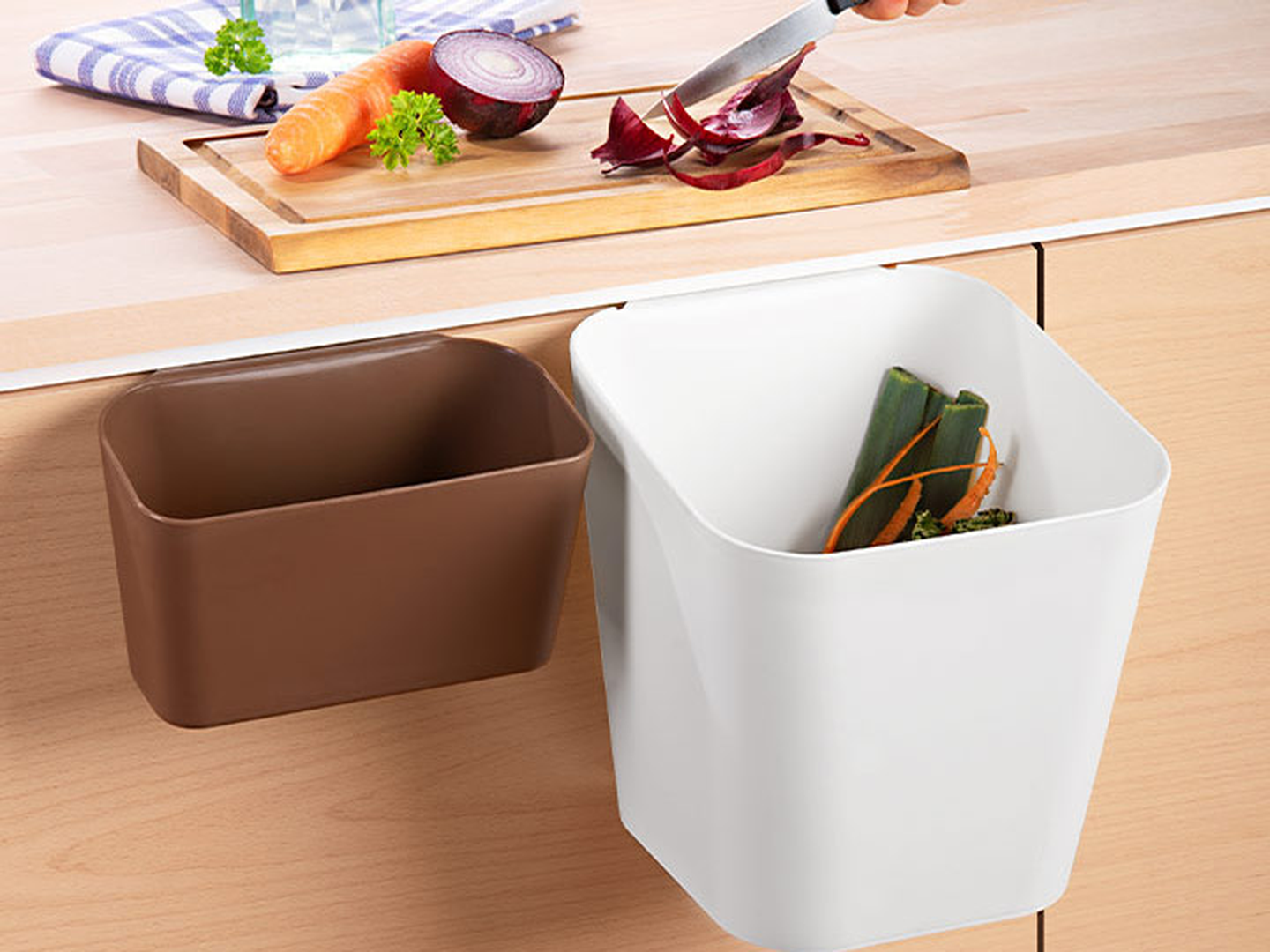 Eine Person schneidet Gemüse auf einem Schneidebrett über einer Küchentheke, an der zwei Mülleimer hängen, ein weißer und ein brauner.