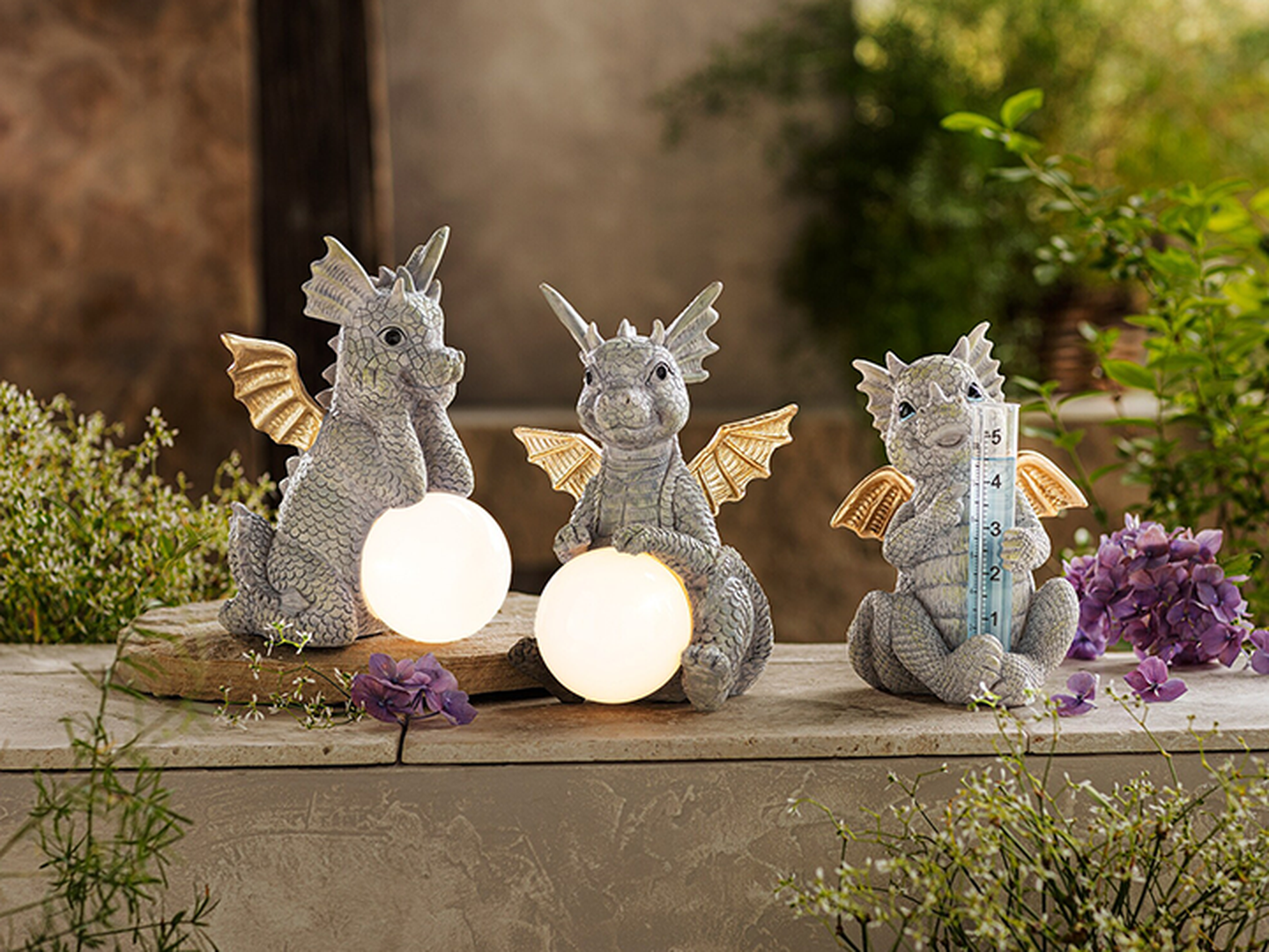 Drei Drachenfiguren mit goldenen Flügeln – zwei halten leuchtende Kugeln, eine hält ein Thermometer –, umgeben von Blumen und Grünpflanzen.