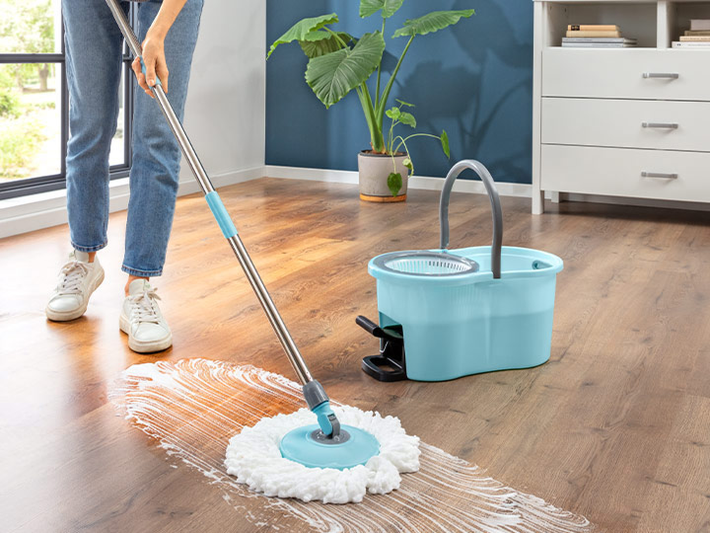Eine Person wischt mit einem blauen Mopp und Eimer einen Holzboden in einem modernen Zimmer mit Pflanzen und einer Kommode.