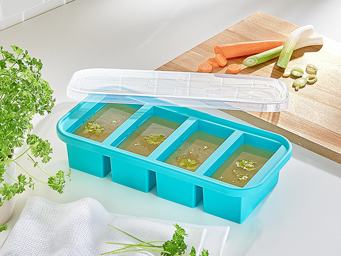 Eine blaue Silikon-Eiswürfelform mit Fächern, die mit Brühe und Kräutern gefüllt sind; sie steht auf einer Arbeitsplatte neben frischem Gemüse und einem weißen Tuch.
