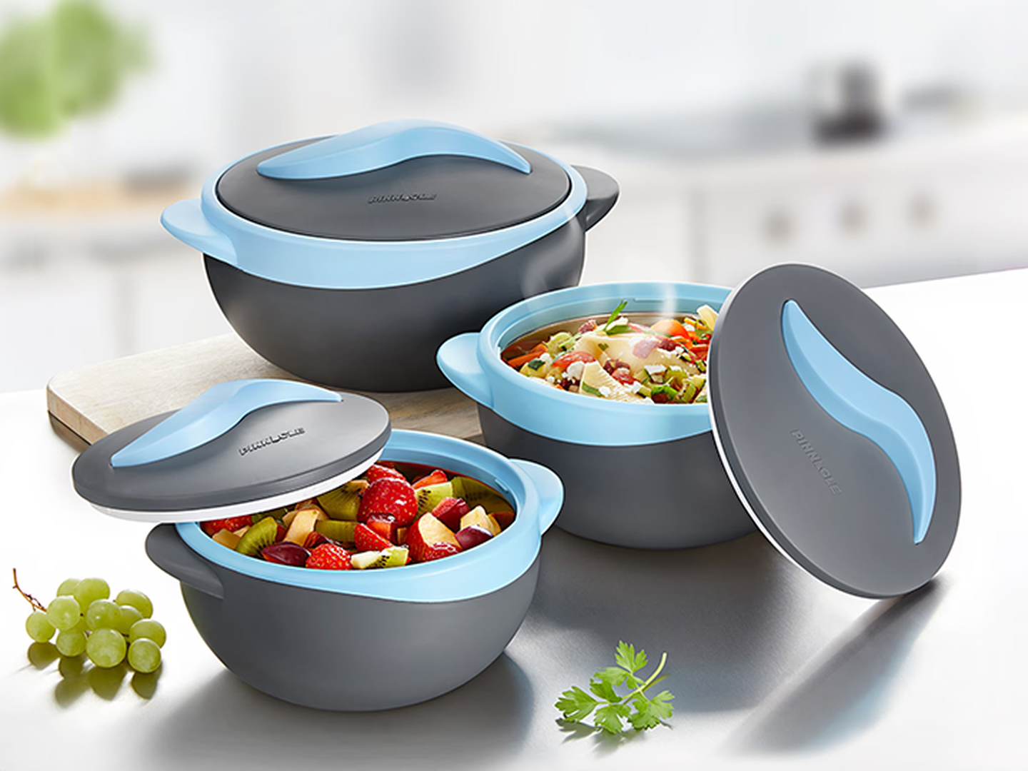 Drei graue Schüsseln mit blauen Deckeln stehen auf einer Arbeitsplatte, gefüllt mit Obstsalat und gemischtem Gemüse. Ein Zweig Petersilie und Weintrauben liegen daneben.