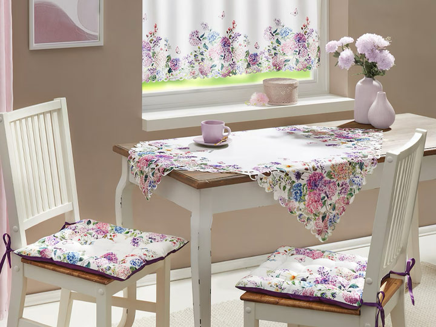 Ein gemütlicher Essbereich mit geblümten Kissen und Tischdecke, einer Teetasse auf dem Tisch und zartrosa Blumen am Fenster mit geblümten Vorhängen.