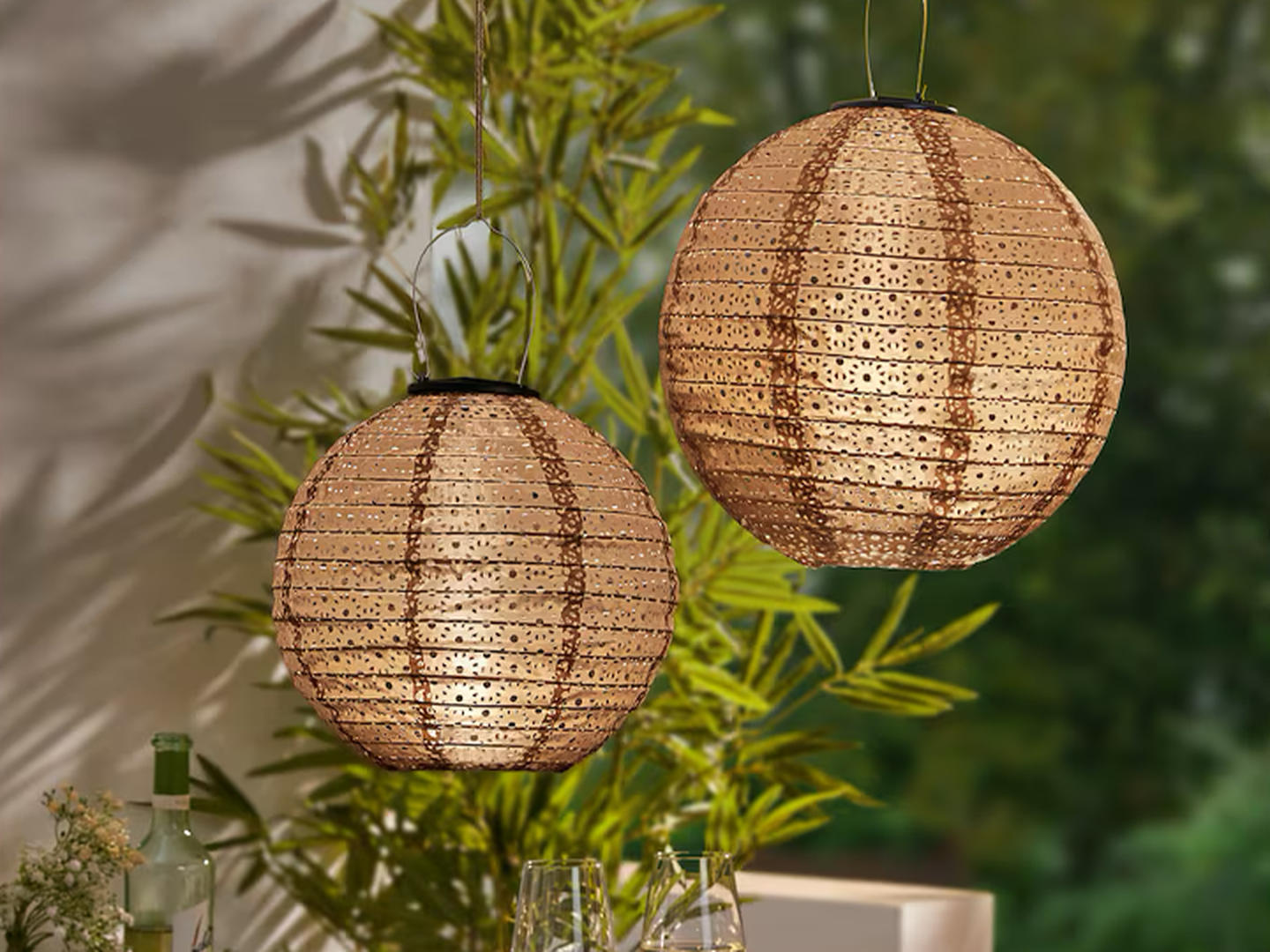 Zwei strukturierte Papierlaternen hängen im Freien, im Hintergrund sind Pflanzen und Schatten zu sehen, die eine warme und einladende Atmosphäre schaffen.