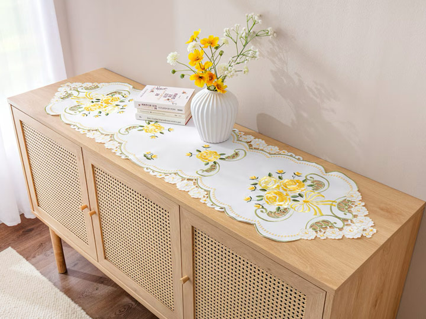 Ein hölzernes Sideboard mit einem floralen Tischläufer, einer weißen Vase mit gelben Blumen und einem kleinen Buch darauf in einem hellen Raum.
