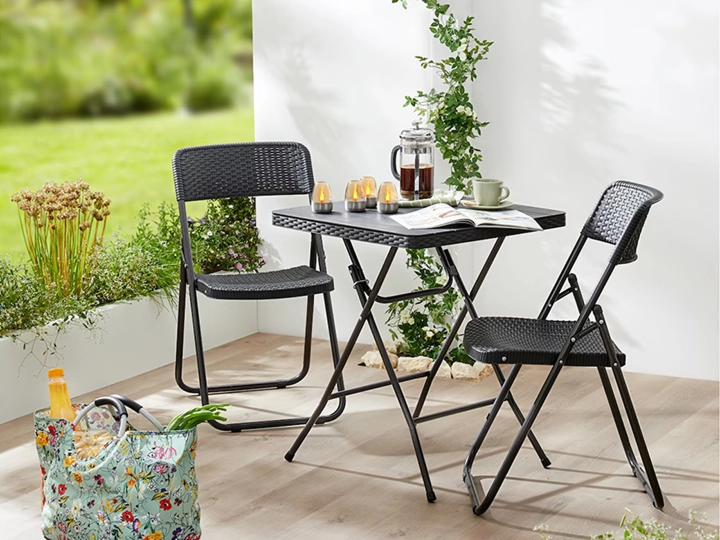 Sitzgruppe mit einem schwarzen Korbtisch und zwei Stühlen, Kerzen, einem Kaffeeservice und einer geblümten Tasche.