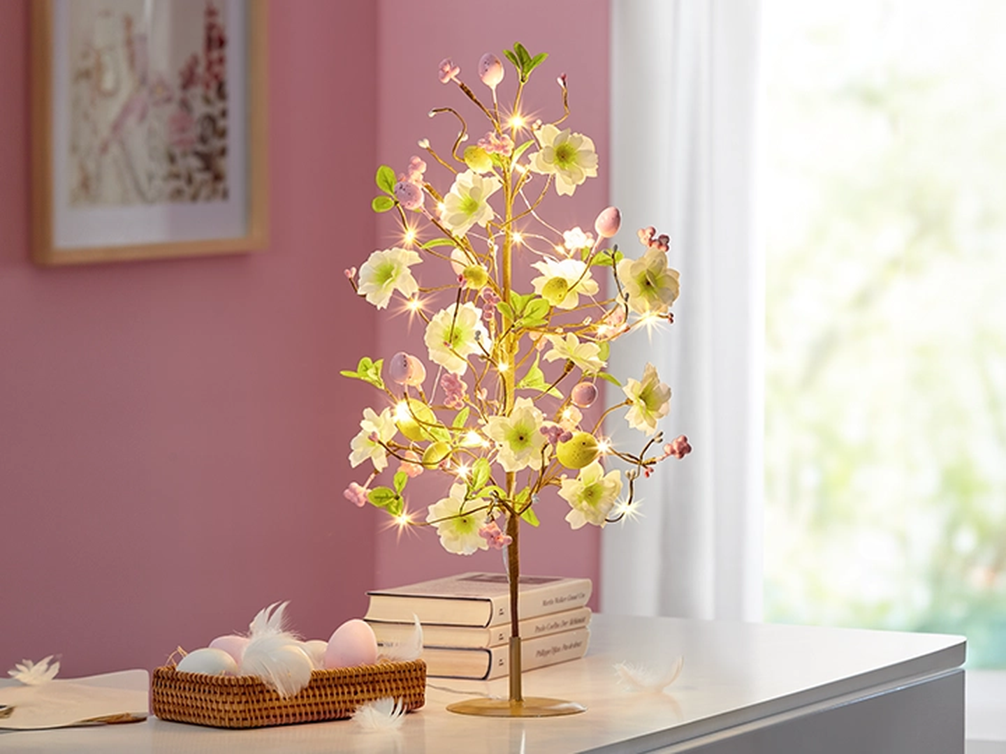 Beleuchtete Blumenbaumlampe mit leuchtenden Blüten auf einem weißen Tisch, neben Büchern und einem dekorativen Korb mit pastellfarbenen Eiern in einem rosa Zimmer.