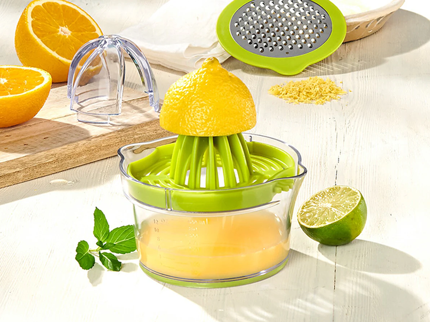 Eine Zitruspresse mit einer halben Zitrone, frischem Saft, Orangenhälften und einer Limette auf einem Holzbrett. Reibe und Zitronenschale daneben.