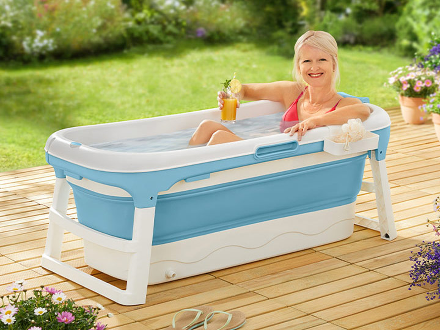 Eine Frau entspannt sich im Freien in einer tragbaren Badewanne, hält ein Getränk in der Hand und steht umgeben von Grün und Blumen auf einer Holzterrasse.