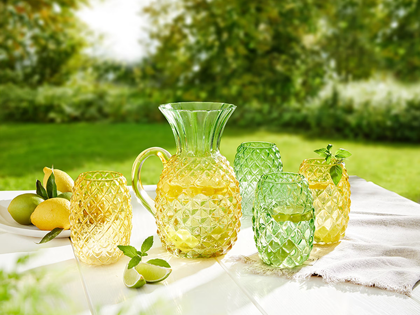 Ein farbenfroher Glaskrug und Gläser stehen auf einem Tisch, gefüllt mit Limonade, umgeben von Zitronen und Minze, vor der Kulisse eines üppig grünen Gartens.