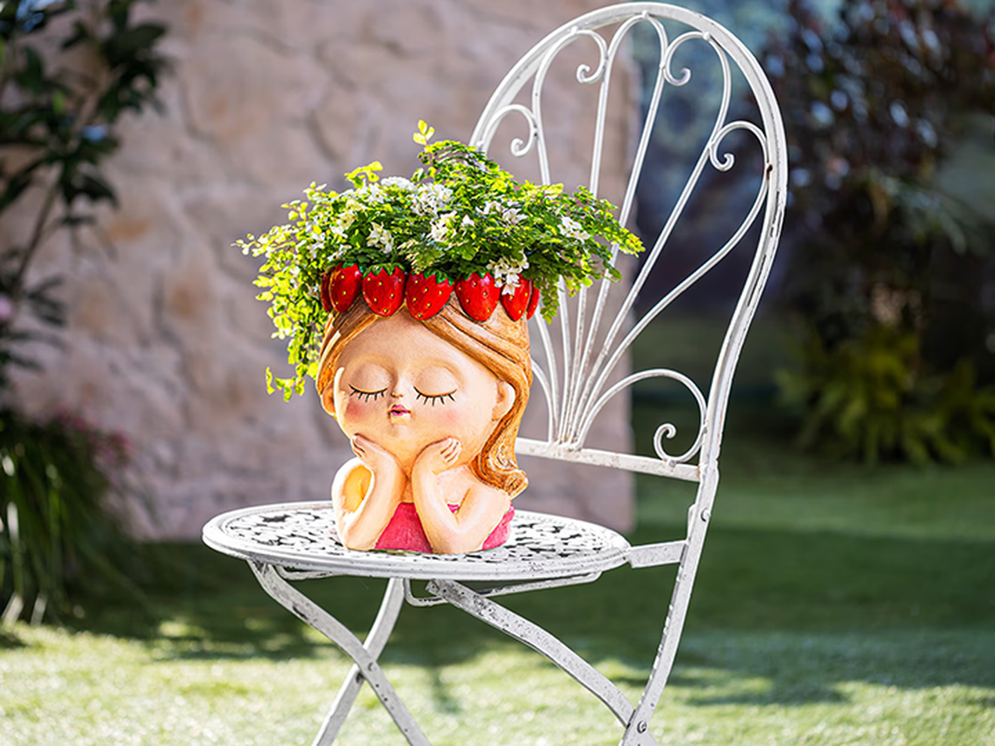 Ein verspielter Pflanzkübel in Form eines Mädchenkopfes, verziert mit Blattpflanzen und Erdbeermotiven, steht auf einem eleganten Metallstuhl in einem Garten.