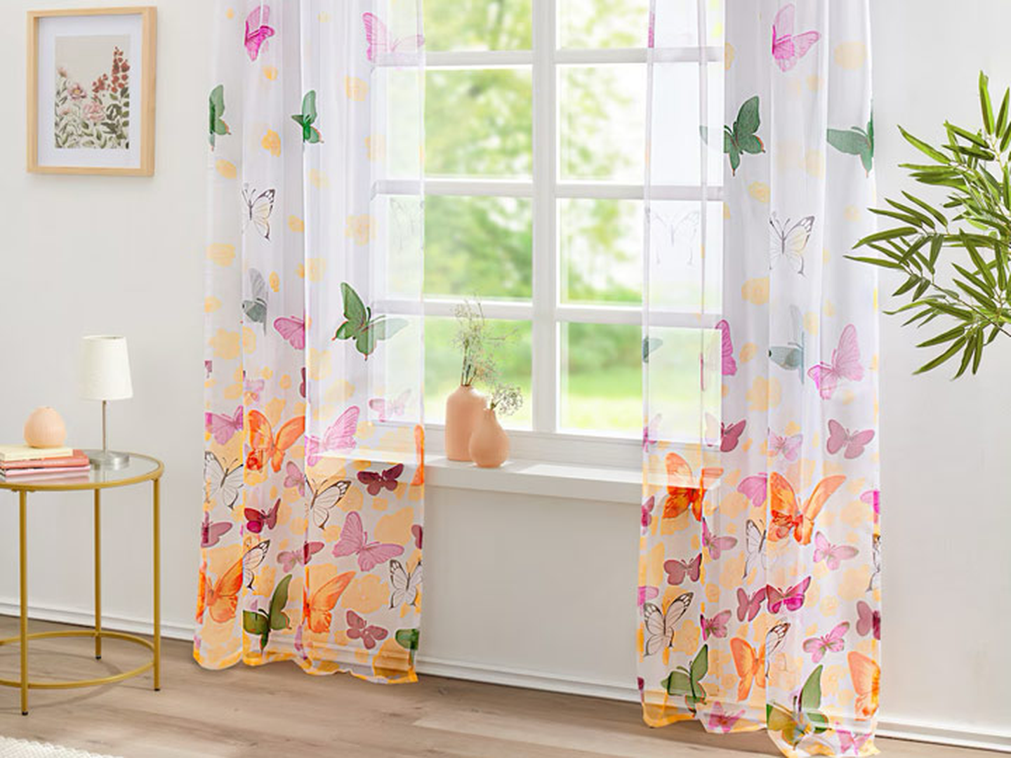 Vor einem Fenster hängen hauchzarte Vorhänge mit bunten Schmetterlingen und Blumen. Ein kleiner Tisch und eine Pflanze stehen daneben, und draußen ist Grün zu sehen.