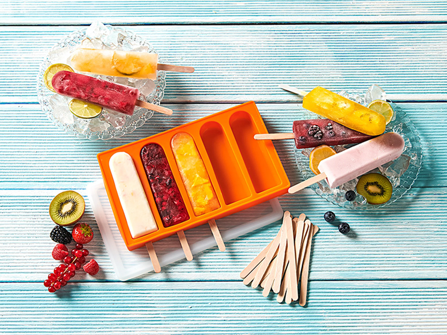 Bunte, selbstgemachte Eis am Stiel in einer orangefarbenen Form, mit verschiedenen Früchten, auf einem blauen Holztisch.