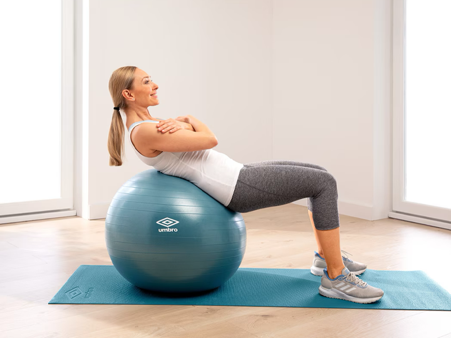 Eine Frau, die auf einem blauen Gymnastikball trainiert, trägt ein weißes Tanktop und graue Leggings und hat die Arme vor der Brust verschränkt. Sie befindet sich auf einer blauen Matte in einem Innenraum.