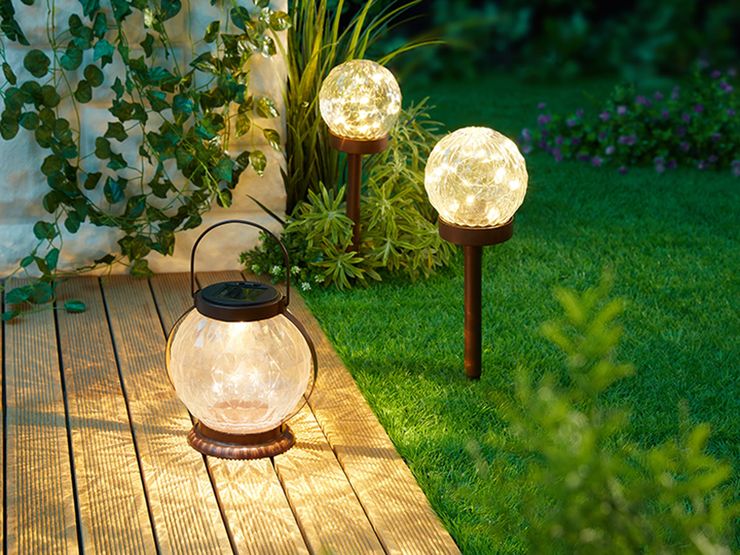 Im Garten leuchtende Solarleuchten, umgeben von grünem Gras und Pflanzen. Eine Laterne steht auf einer Holzterrasse neben zwei kugelförmigen Gartenleuchten.