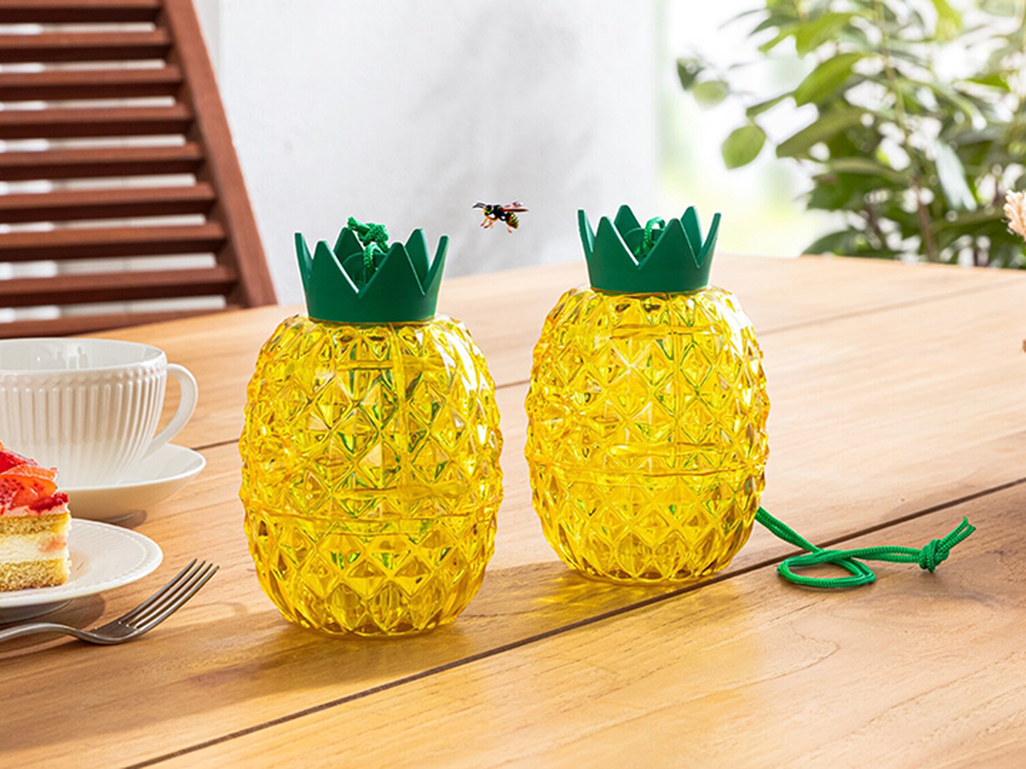 Zwei gelbe, an Ananas erinnernde Behälter mit grünen Deckeln stehen auf einem Holztisch, in der Nähe eines Tellers mit Kuchen und einer Tasse; eine Biene schwebt darüber.