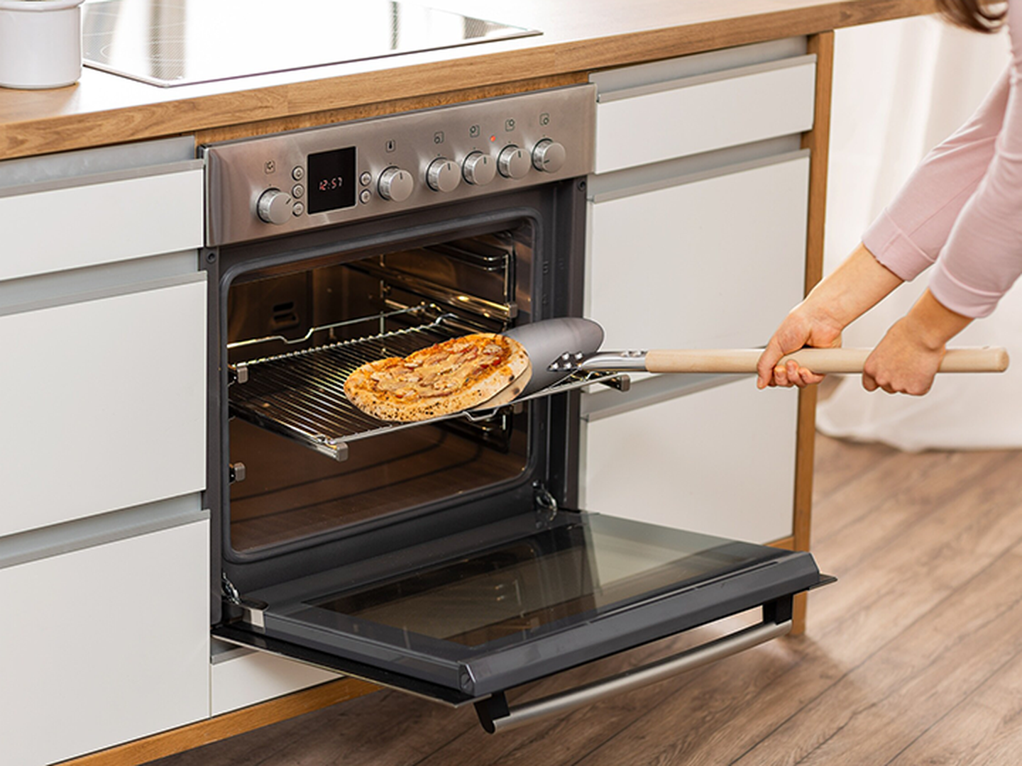 Eine Person nimmt in einer modernen Küche mit einem Pizzaschieber eine Pizza aus einem offenen Backofen.