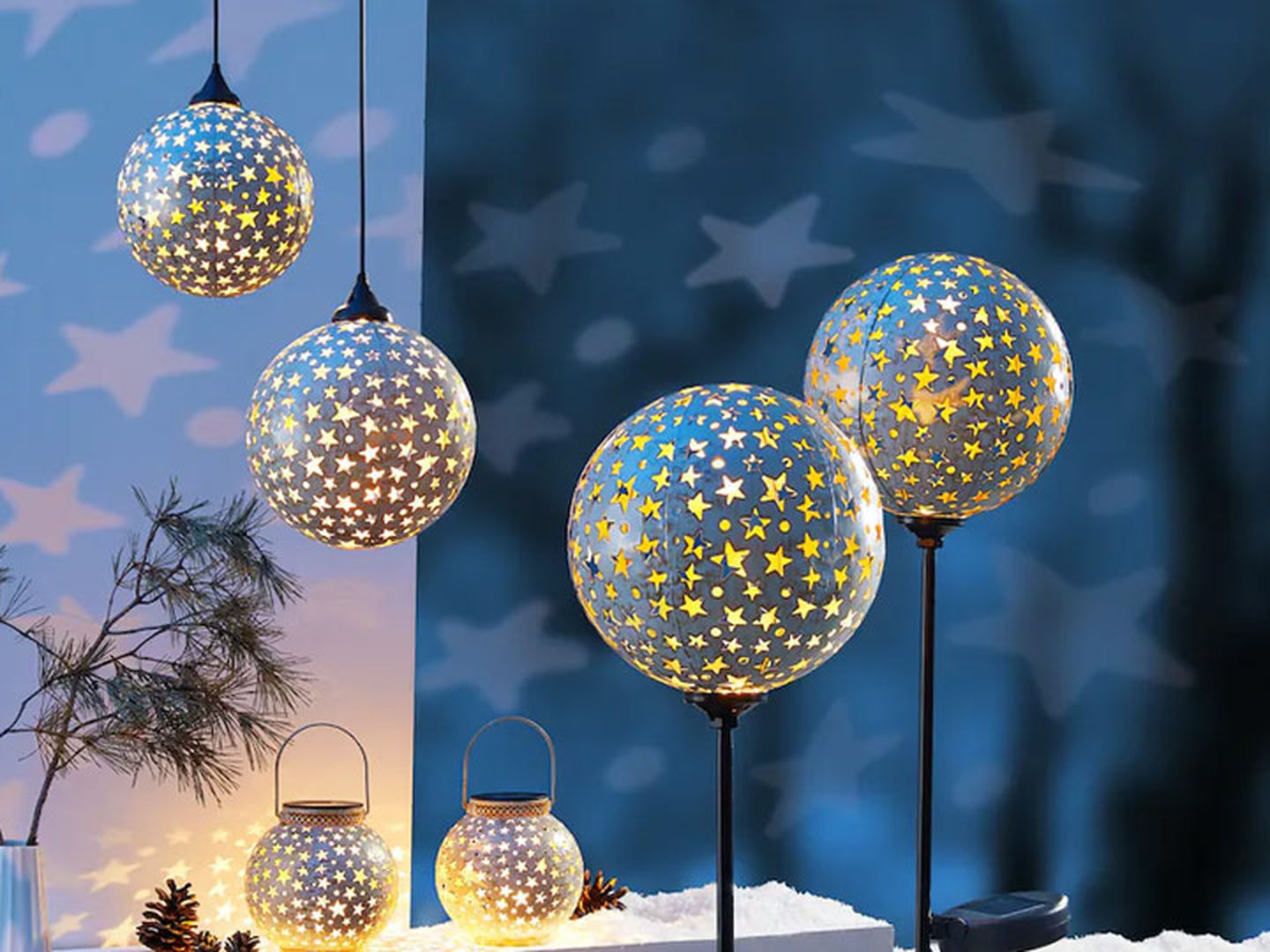 Leuchtende, kugelförmige Laternen mit Sternenmuster hängen und stehen auf Ständern und werfen sternförmige Schatten auf eine blaue Wand über einer schneebedeckten Tischplatte mit Tannenzapfen.