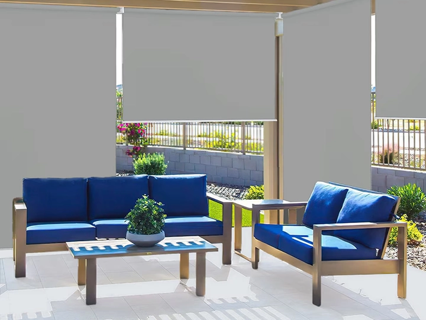 Terrasse mit blauen Polstermöbeln, einem Tisch mit Pflanze unter einer Pergola mit Sonnenschutz. Garten und Zaun im Hintergrund.