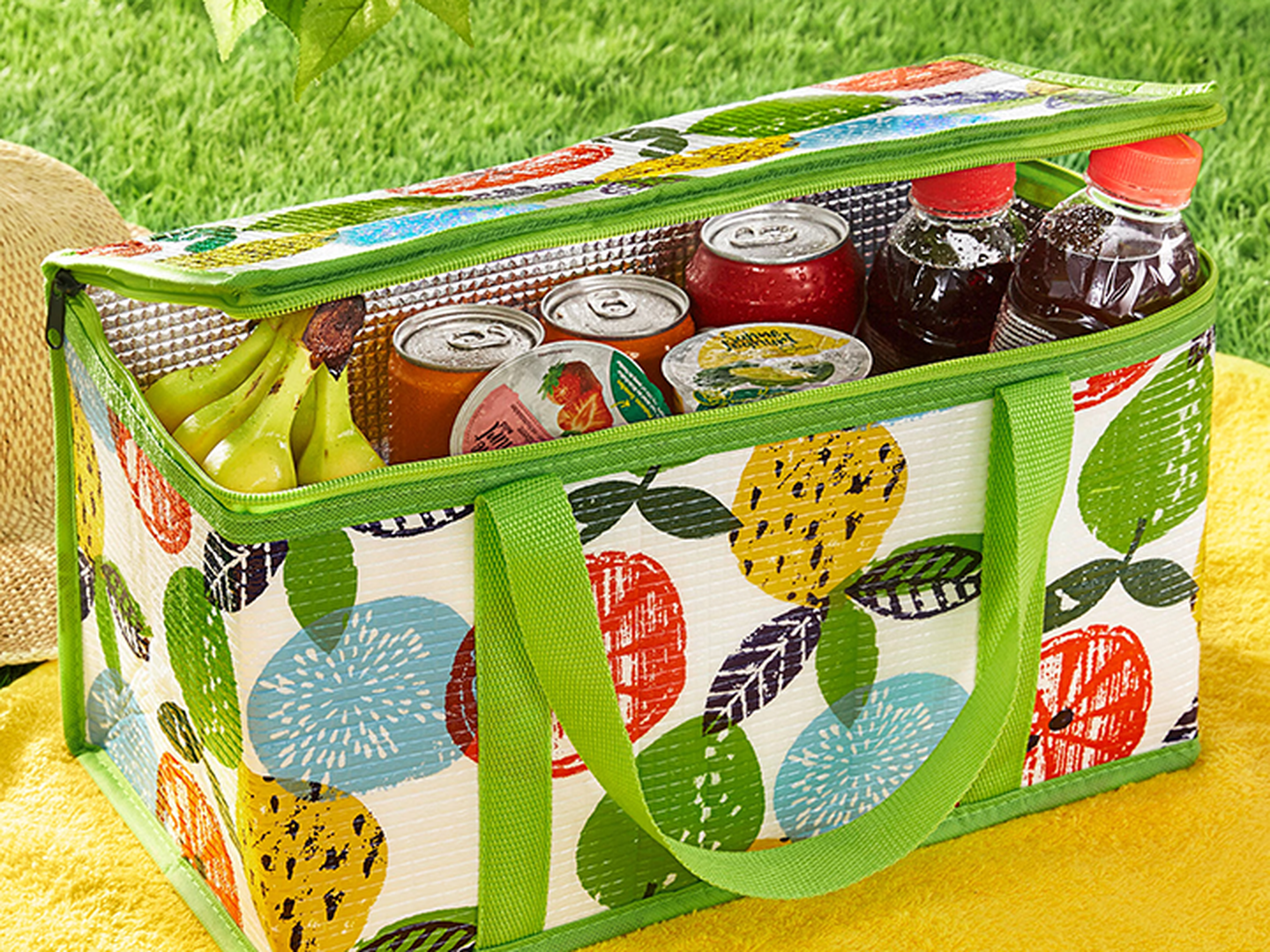 Bunte Kühltasche mit Fruchtmuster, gefüllt mit Bananen sowie Getränken in Dosen und Flaschen, auf einer gelben Decke im Gras.
