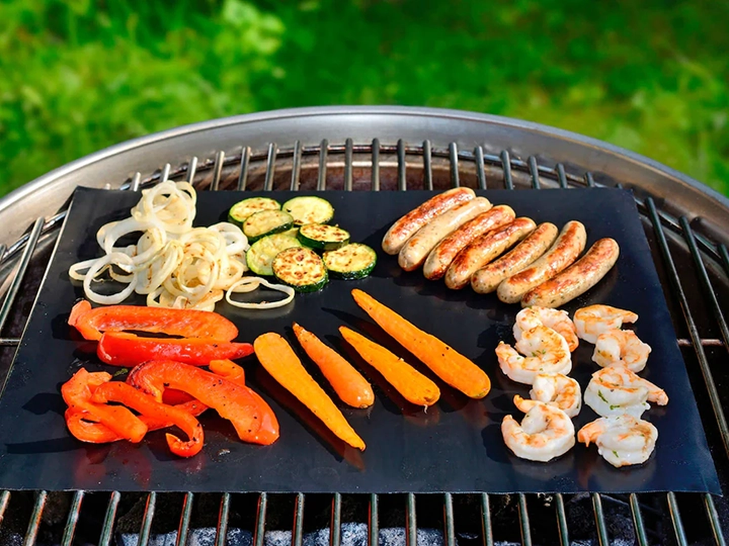 Verschiedene Gemüsesorten, Würstchen und Garnelen brutzeln auf einer Grillmatte, im Hintergrund ist ein grüner Rasen zu sehen.