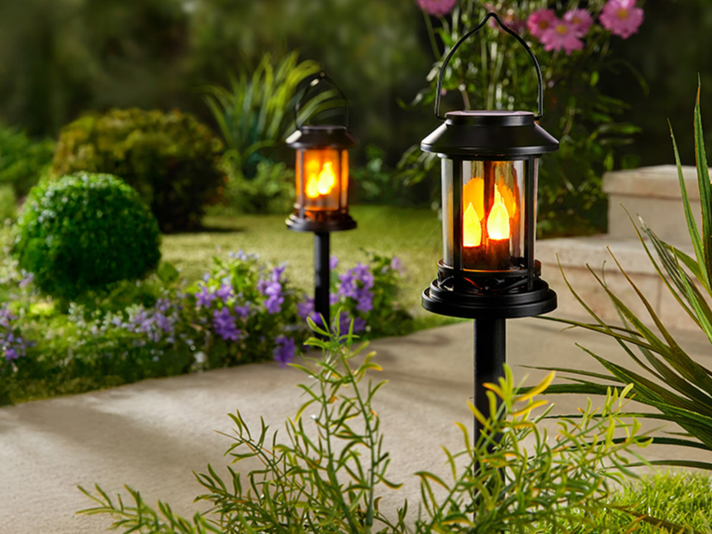Schwarze Laternen beleuchten einen Gartenweg mit flackerndem Flammenschein, umgeben von üppigem Grün und violetten Blumen.
