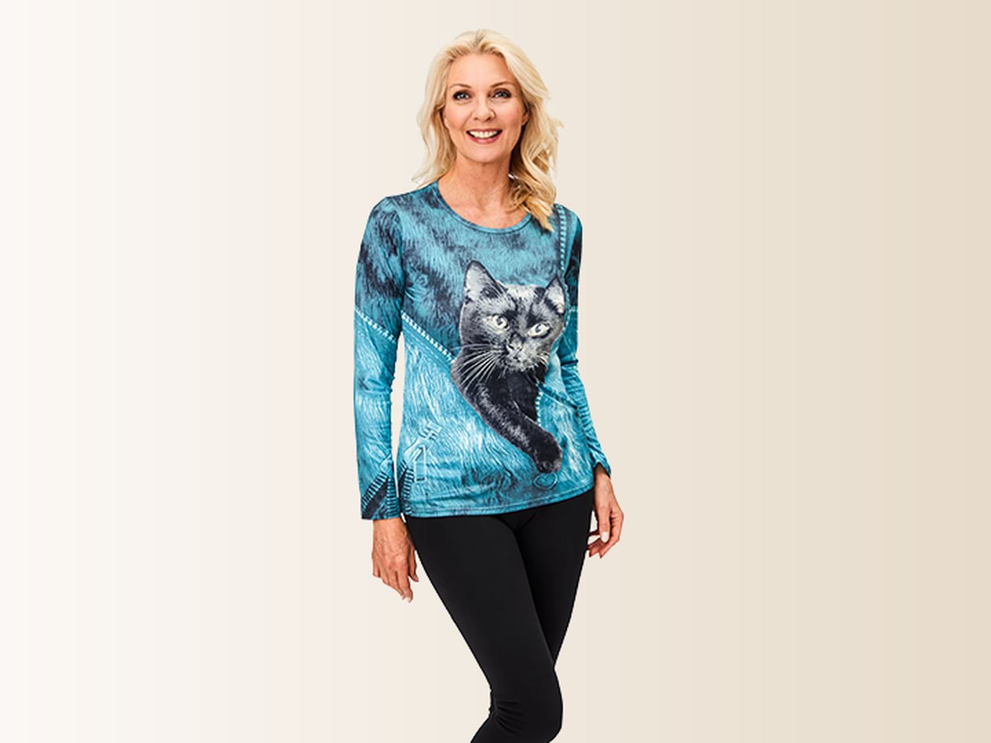 Eine lächelnde Frau in einem türkisfarbenen Pullover mit Katzenmotiv und schwarzer Hose, die vor einem hellen Hintergrund steht.