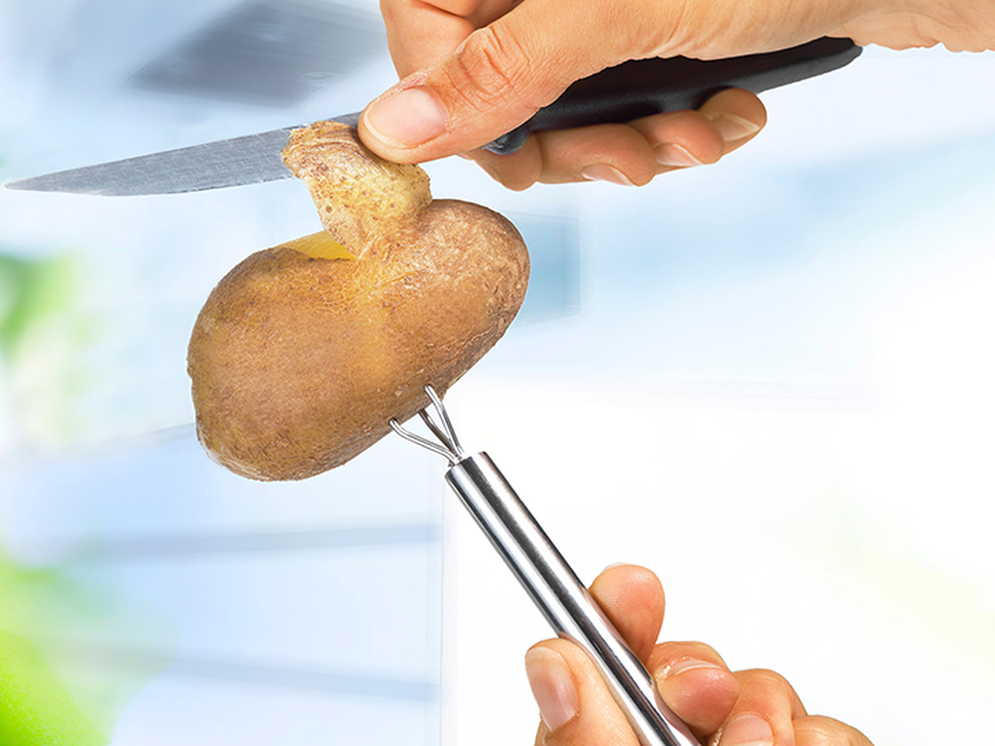Eine Hand schält eine Kartoffel mit einem Messer, während sie diese mit einem Kartoffelhalter aus Edelstahl fixiert. Der Hintergrund ist unscharf.