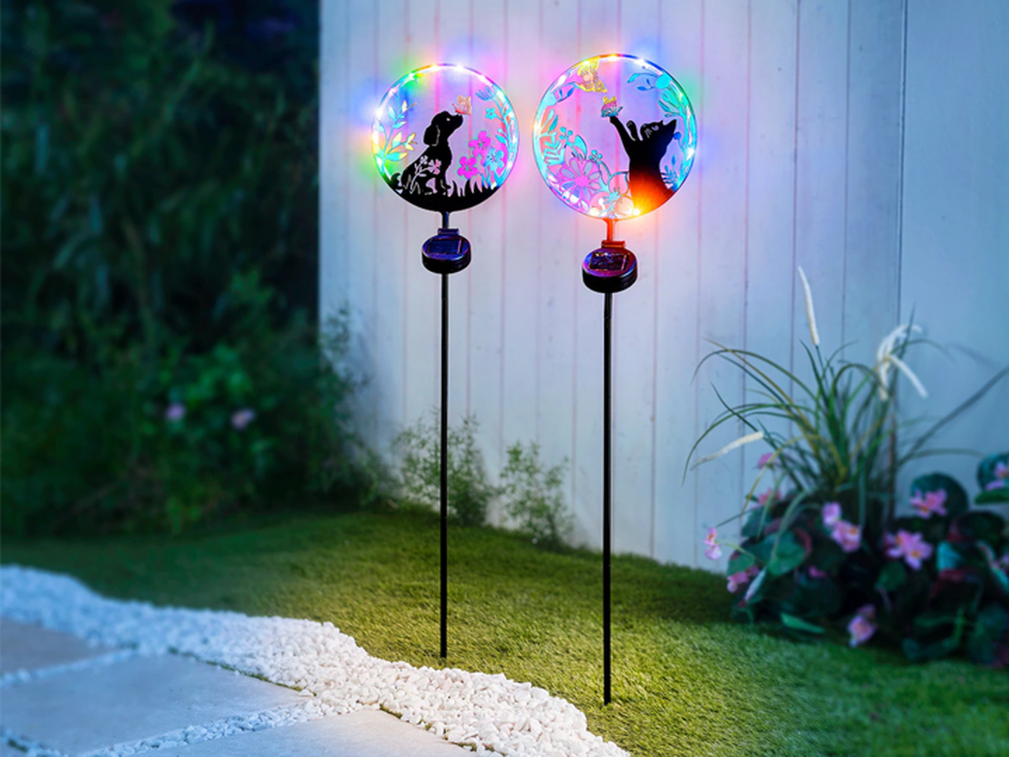 Zwei bunte Gartenleuchten mit den Silhouetten eines Hundes und einer Katze auf einer Rasenfläche in der Nähe eines weißen Zauns und blühender Pflanzen.