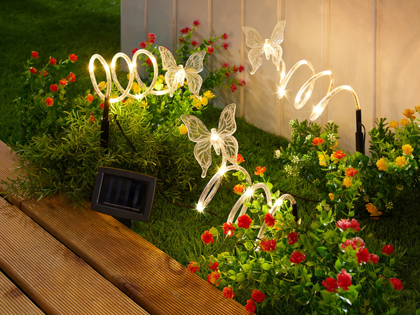 Schmetterlingsförmige Gartenleuchten erstrahlen zwischen grünen Büschen und bunten Blumen neben einem Holzweg. Das Solarpanel ist im Gras sichtbar.
