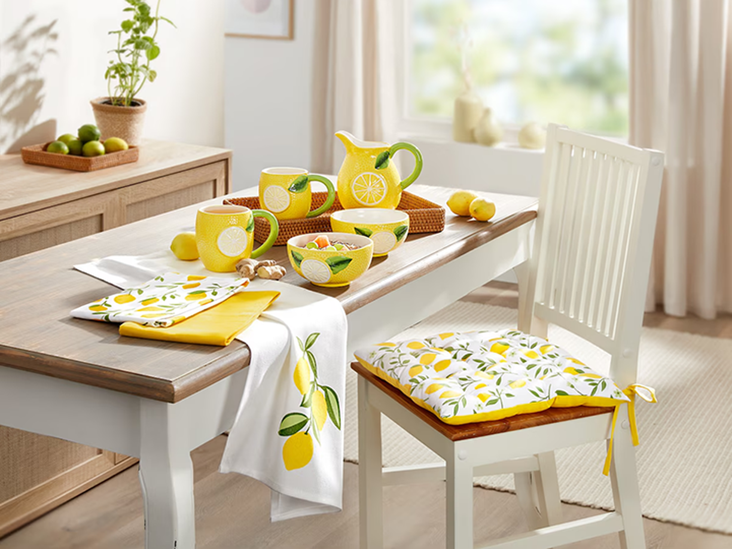 Eine helle Küchenszene mit Dekoration im Zitronen-Design – darunter ein Krug, Tassen, Schalen, Servietten und ein Stuhlkissen – auf einem Holztisch.