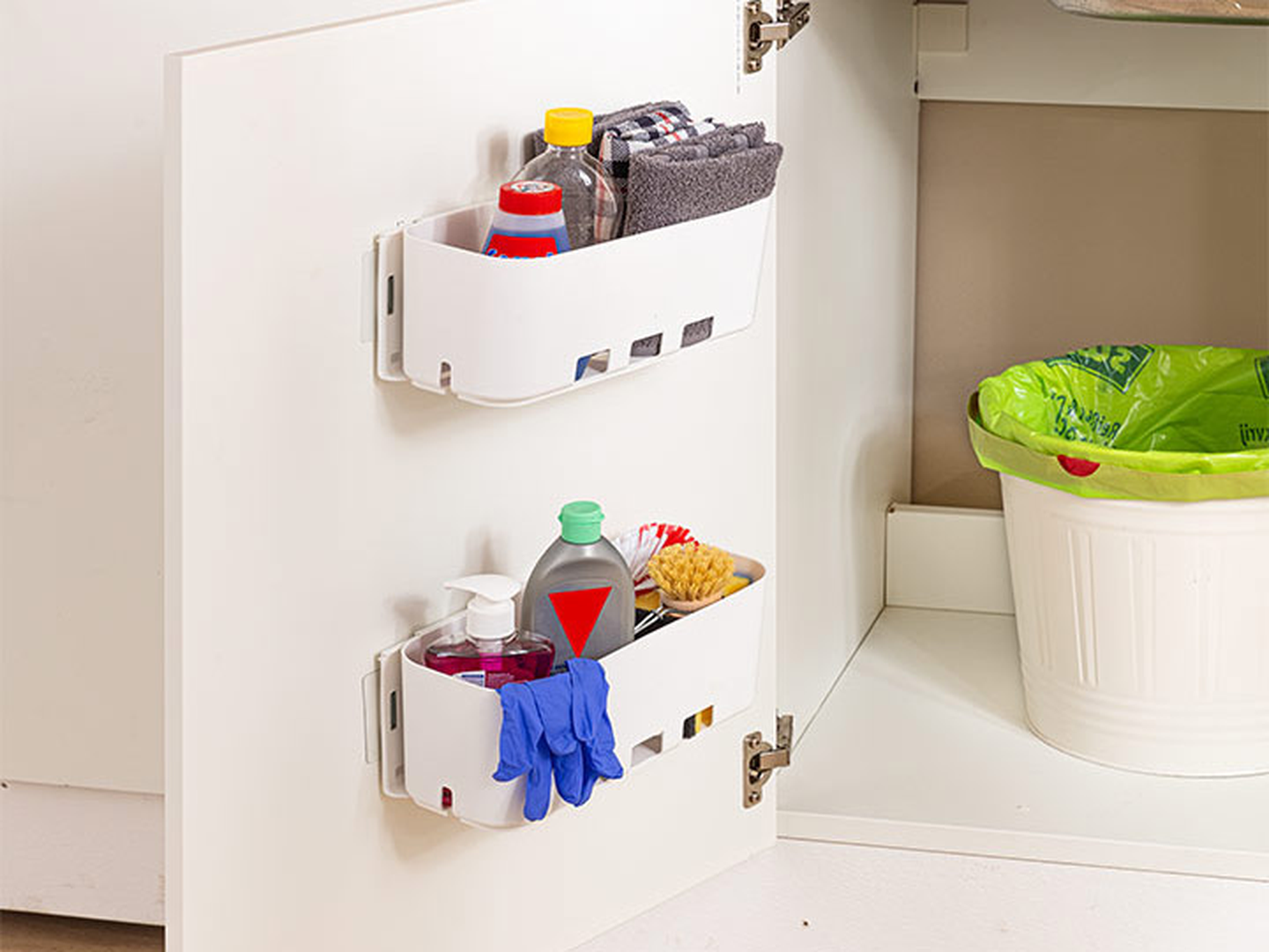Offener Schrank mit zwei weißen Ablagefächern an der Tür, in denen Reinigungsmittel und Handschuhe aufbewahrt werden, daneben ein Mülleimer mit grünem Einsatz.