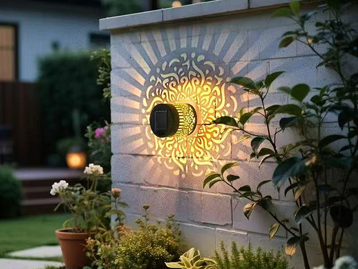 Eine dekorative Wandleuchte wirft in der Dämmerung kunstvolle Muster auf eine Gartenmauer, umgeben von Topfpflanzen.