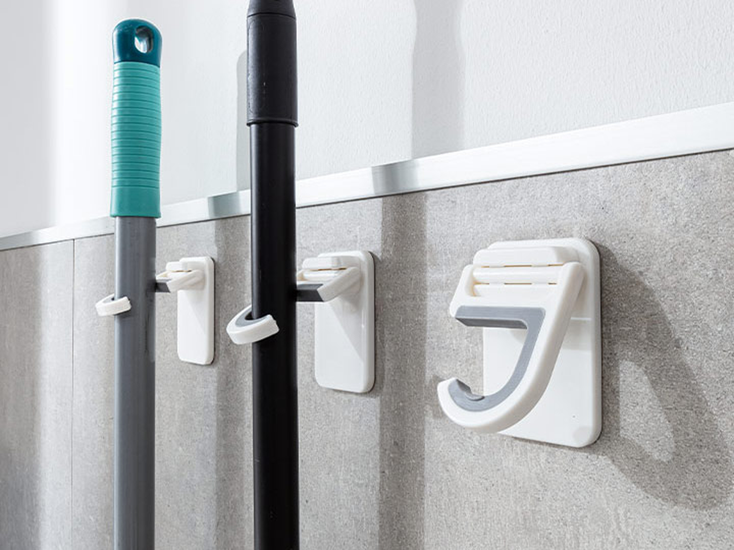 Wandmontierte Halterungen zur Befestigung von Besen und Mopp an einer grau gefliesten Wand, mit Haken zum Aufhängen.