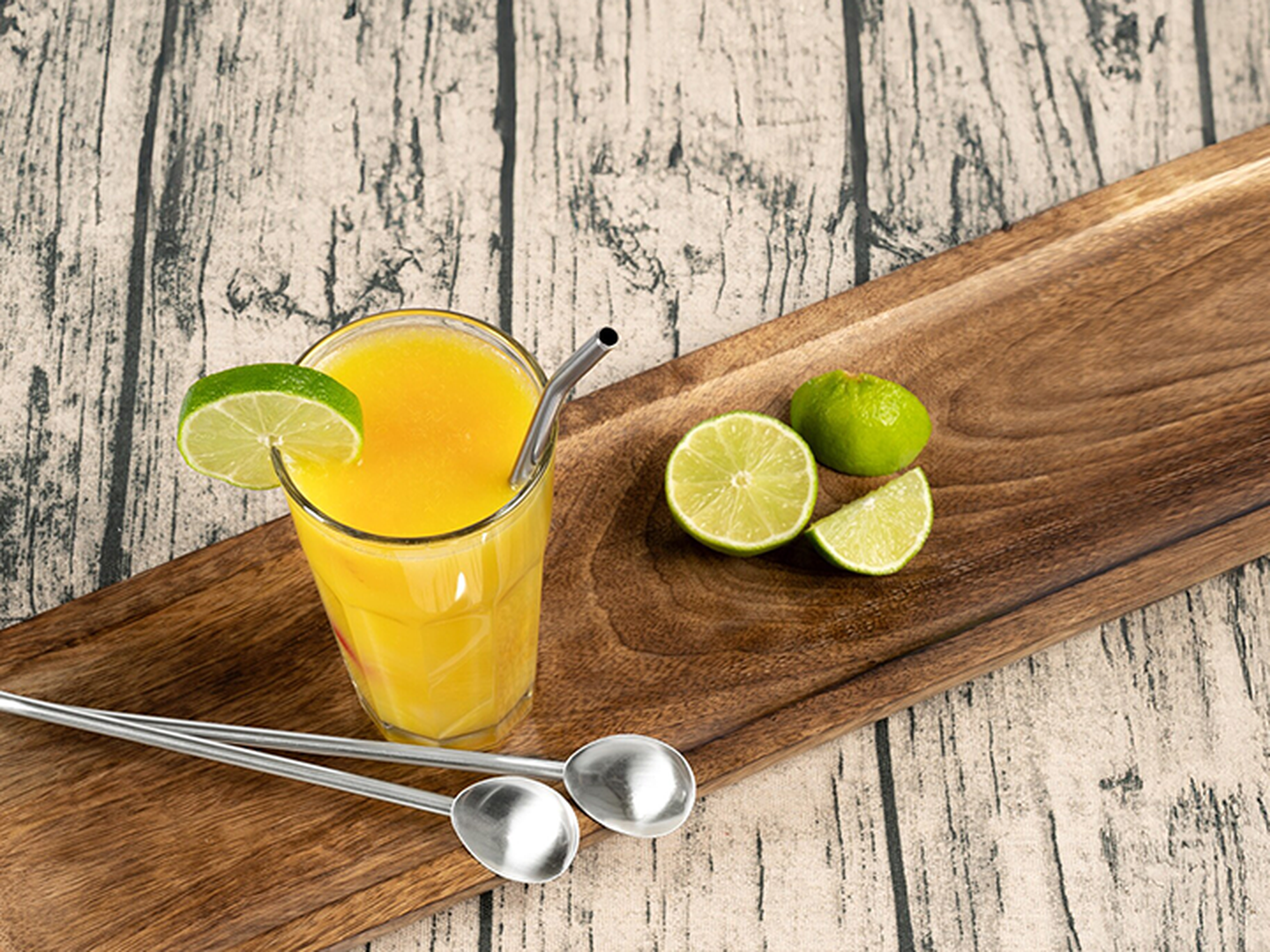 Ein Glas Orangensaft mit einem Limettenstück und einem Metallstrohhalm auf einem Holztablett, begleitet von Limettenscheiben und Metalllöffeln.