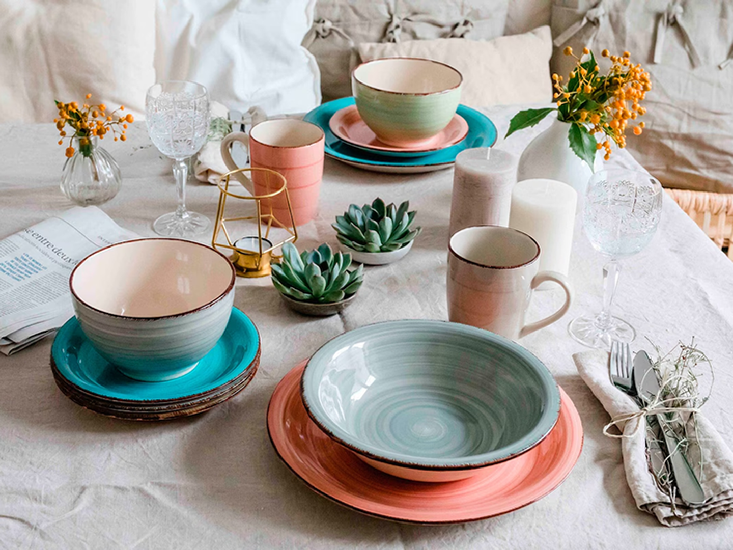 Elegant gedeckter Tisch mit farbenfrohem Geschirr, Gläsern, Kerzen, Sukkulenten und Blumen auf einer Leinentischdecke.