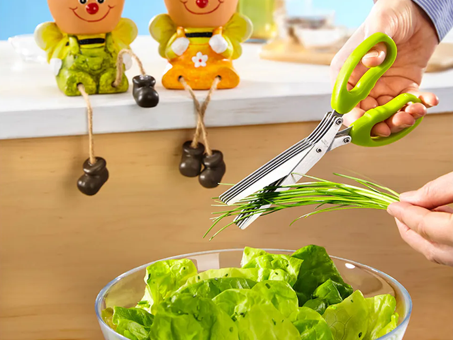 Eine Person schneidet mit einer grünen Mehrklingenschere Kräuter über einer Schüssel mit Salat; zwei dekorative Figuren sitzen daneben.