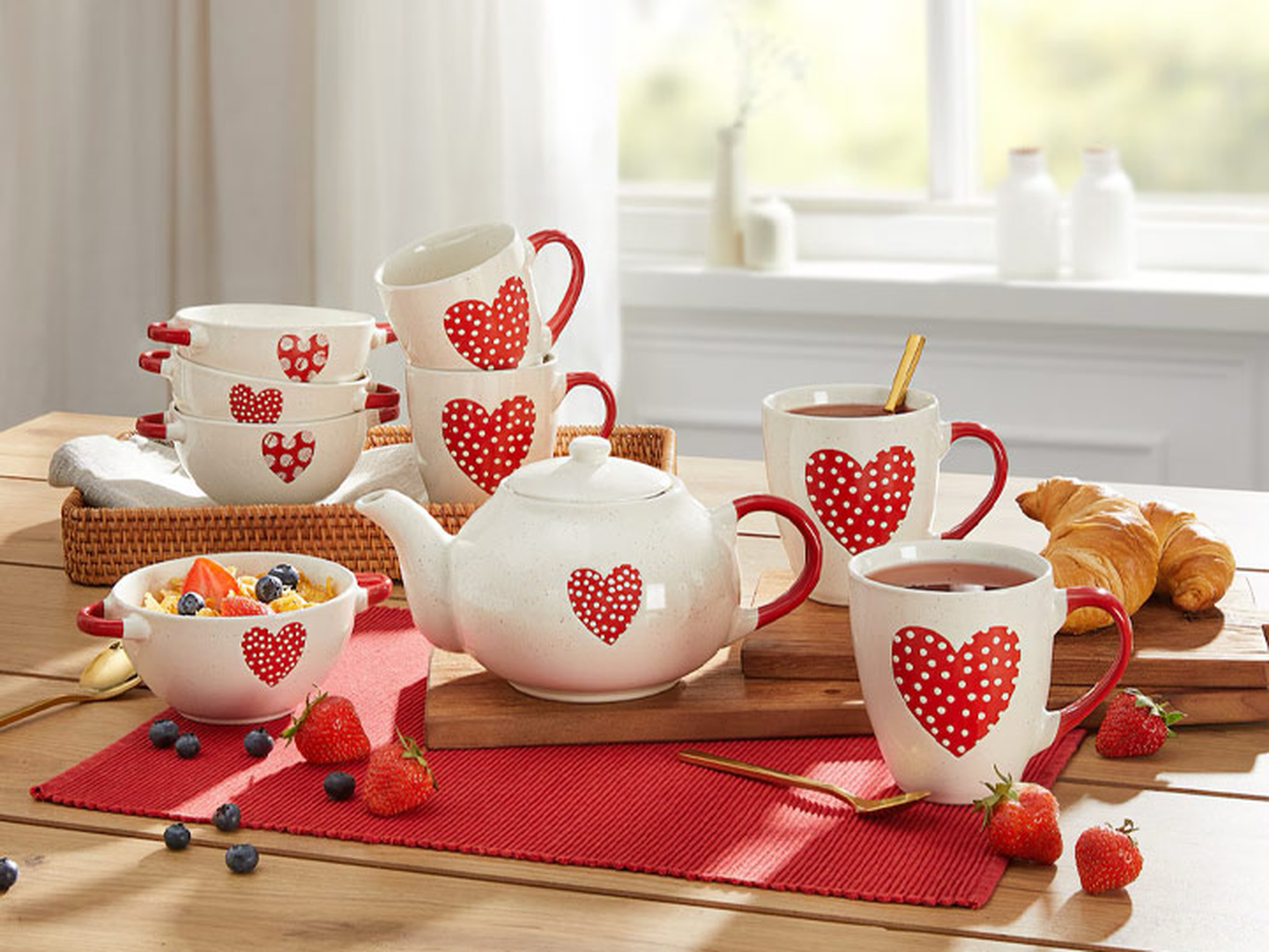 Teeservice mit roten Herzmotiven auf Tassen und Teekanne auf einem Holztisch. Dazu gehören eine Obstschale, Croissants und ein rotes Platzset. Heller Ausblick aus dem Fenster.