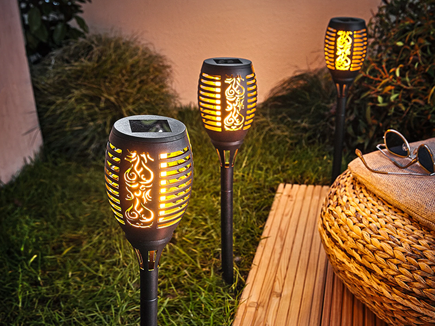 Drei dekorative Solar-Gartenfackeln mit filigranen Mustern verbreiten einen warmen Schein auf einer Rasenfläche, neben einem geflochtenen Korb und einer Holzmatte.