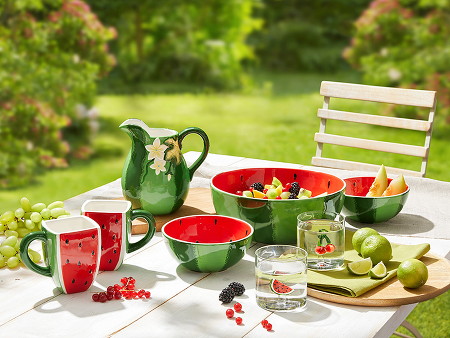 Ein Tisch im Garten, gedeckt mit Schalen, Bechern und Krügen im Wassermelonen-Design – gefüllt mit Obst – sowie Wassergläsern und Limetten.