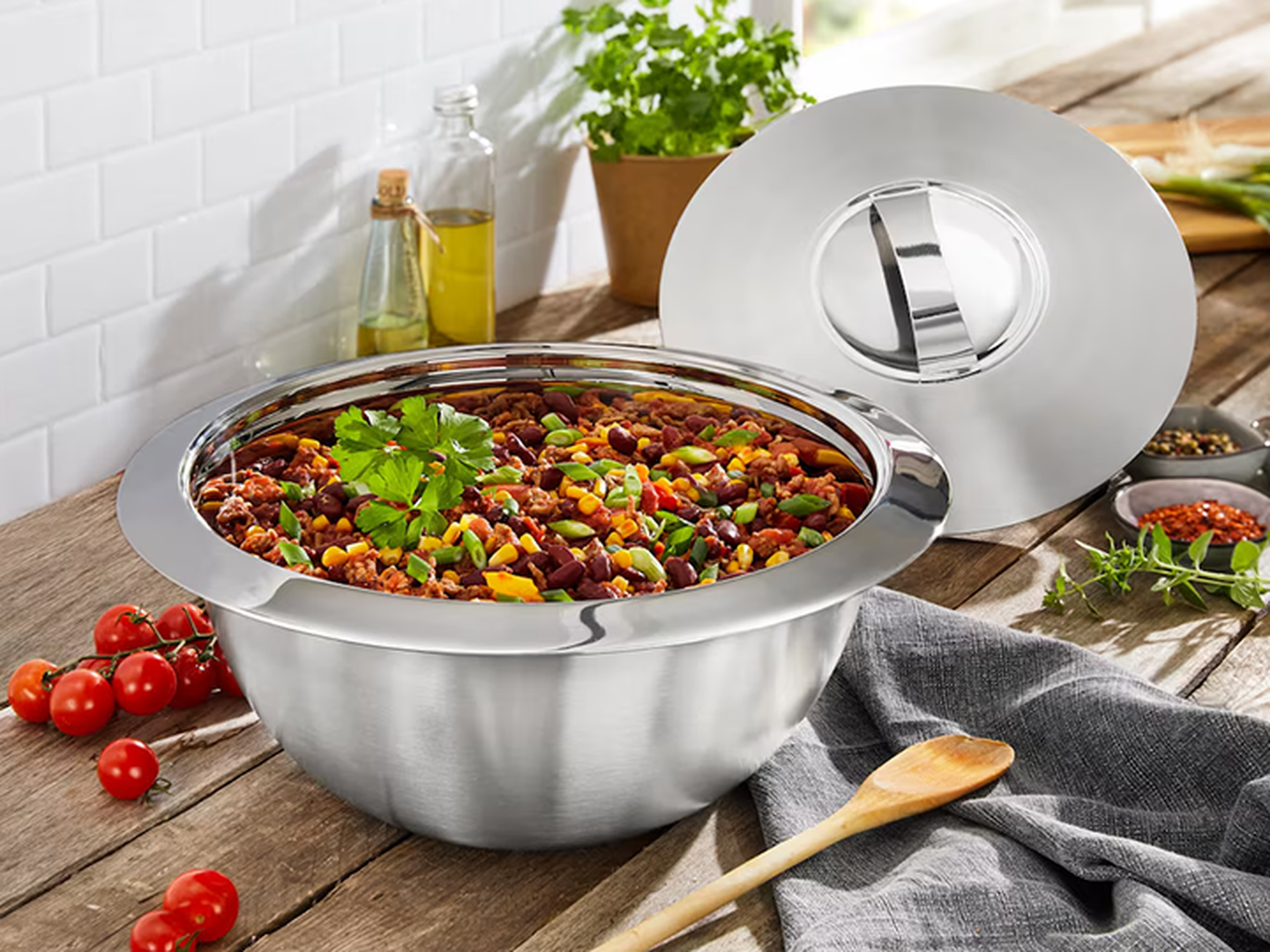 Eine Edelstahlschüssel gefüllt mit Essen, garniert mit Kräutern, daneben ein passender Deckel, Tomaten und Kochutensilien auf einem Holztisch.