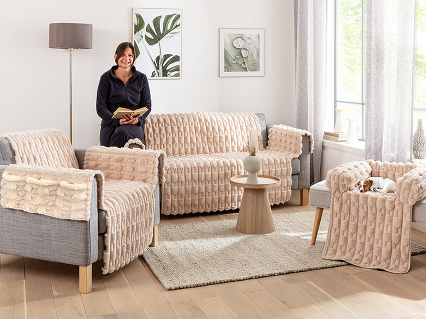Eine Frau steht in ihrem Wohnzimmer mit einem Buch in der Hand, ein Sofa, ein Sessel und ein Hocker mit Hundebett sind mit einem rosa Sofaschoner überzogen