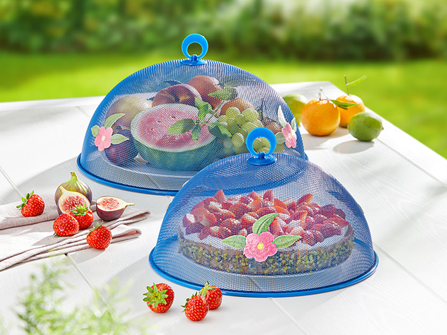 Zwei Netzabdeckungen für Speisen mit blauen Griffen und floralen Verzierungen bedecken verschiedenes Obst und Beeren auf einem weißen Tisch.