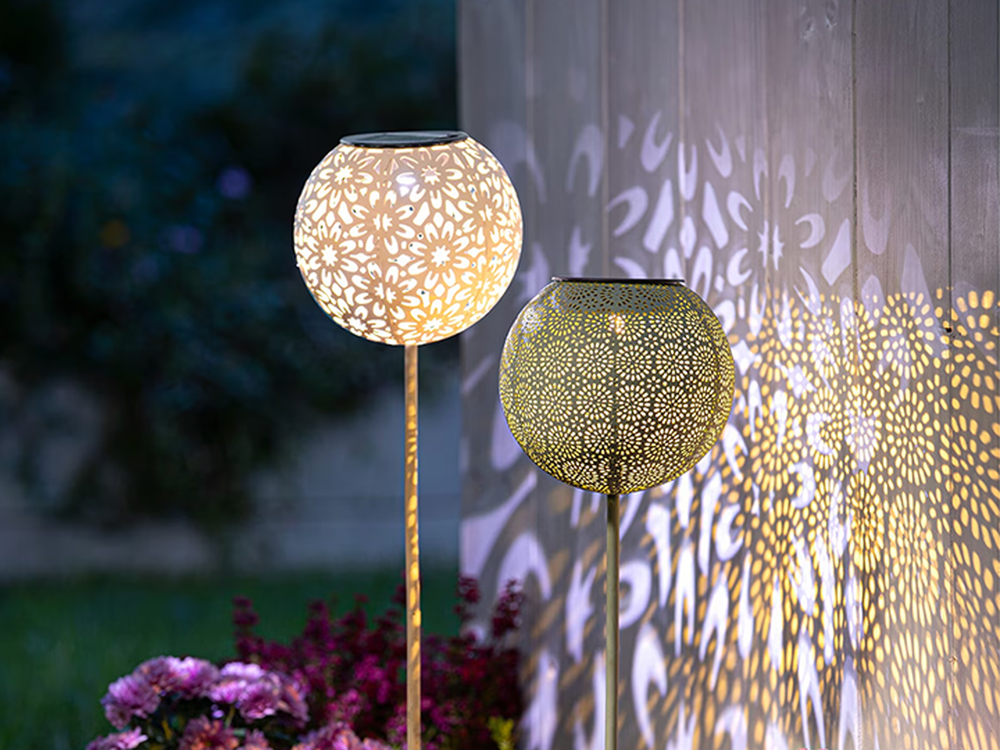 Zwei dekorative, kugelförmige Lampen werfen filigrane Blumenmuster an eine Wand. Eine Lampe leuchtet hell, während die andere ein sanfteres Licht abgibt.