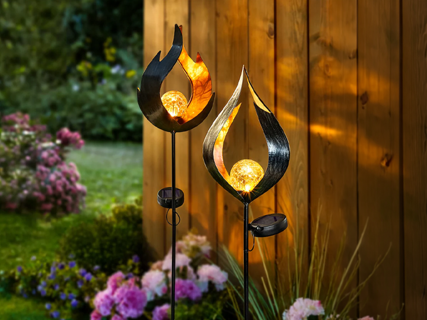 Zwei Solar-Gartenleuchten mit blattförmigen Metallrahmen und leuchtenden Glühbirnen, aufgestellt in einem üppigen Garten in der Nähe eines Holzzauns mit blühenden Blumen.